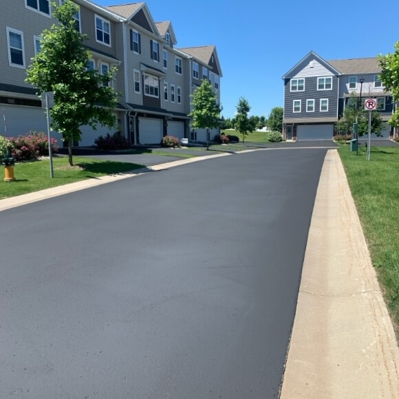 A newly paved asphalt road in a residential neighborhood with townhouses and well-maintained greenery.