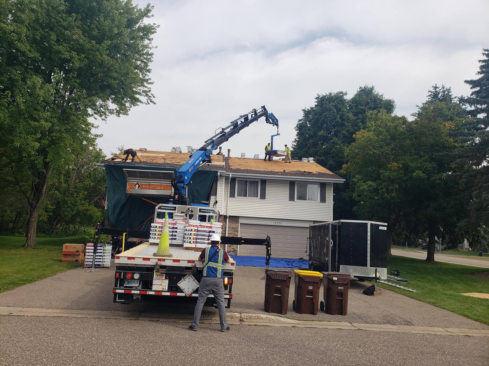 A roofing crew working on a house roof with a crane lifting roofing materials. The yard is filled with roofing supplies and a large vehicle, while workers repair the roof.