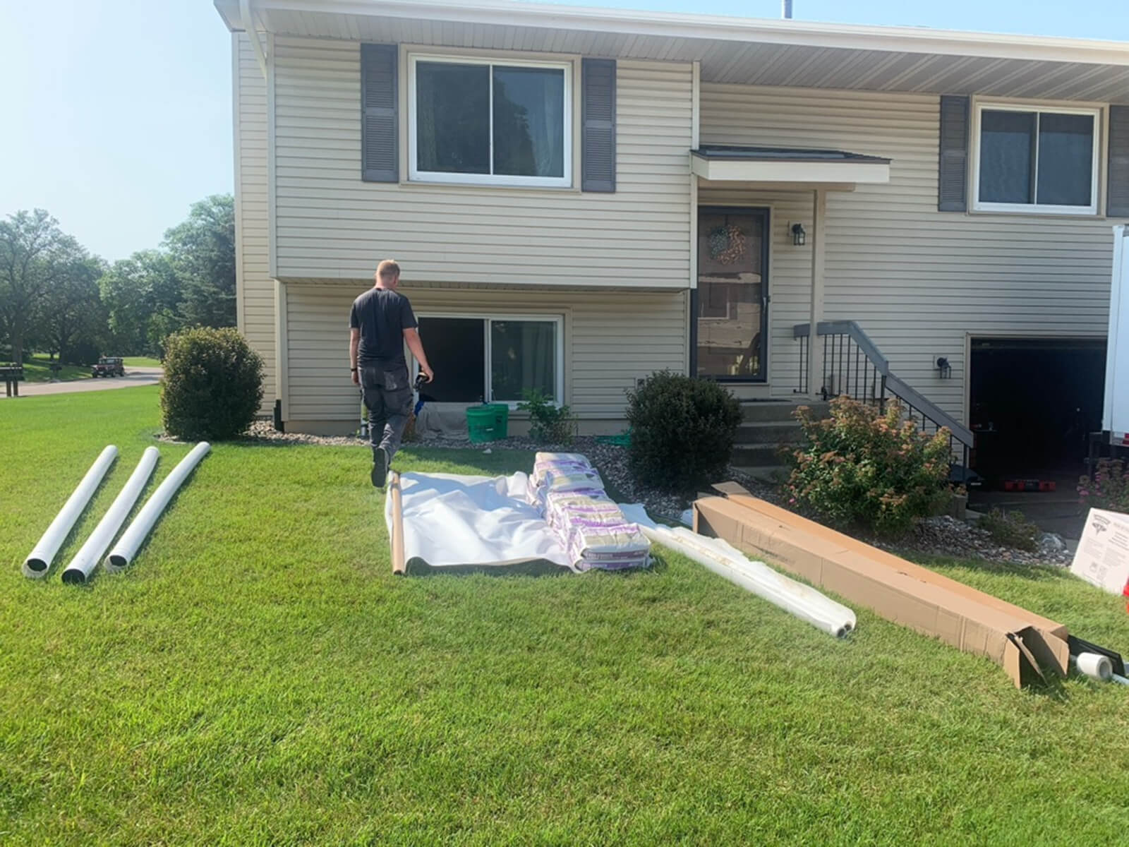 Roofing materials and tools laid out on the lawn in front of a residential home, with a worker walking toward the house, preparing for roof installation.