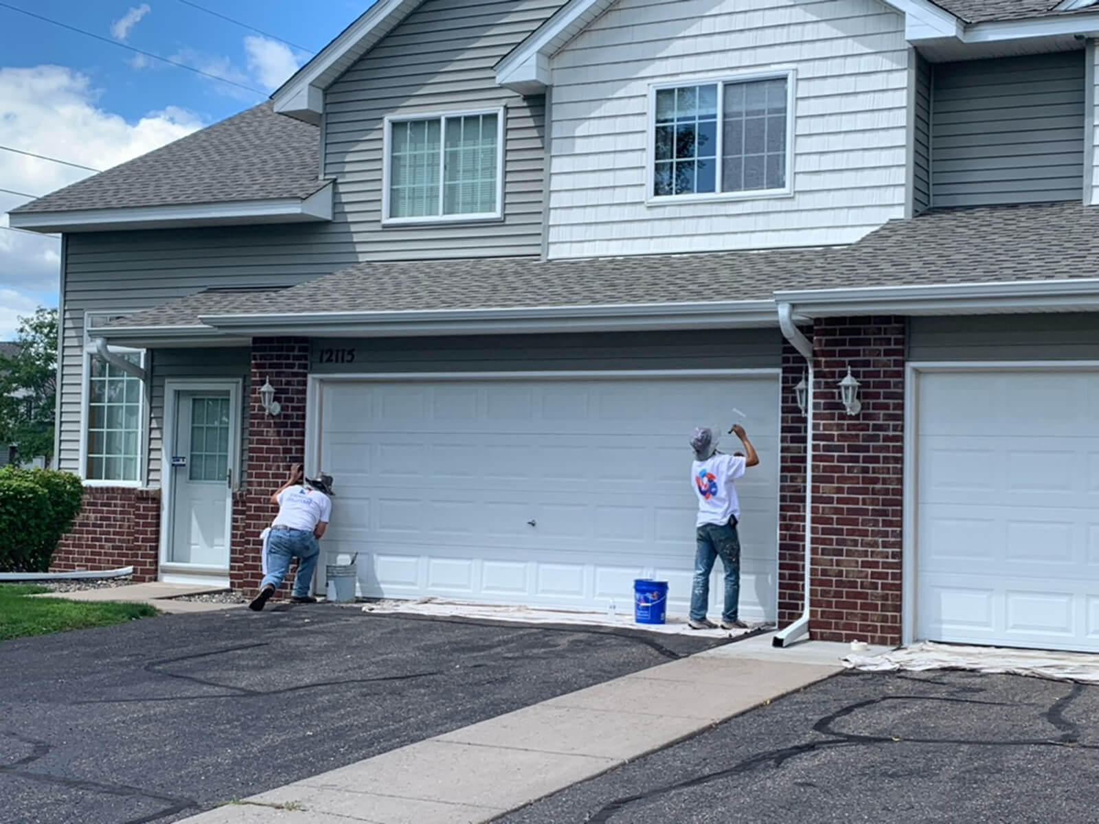 Two painters applying fresh paint to garage doors of a residential home, with drop cloths and paint buckets set up around the area.