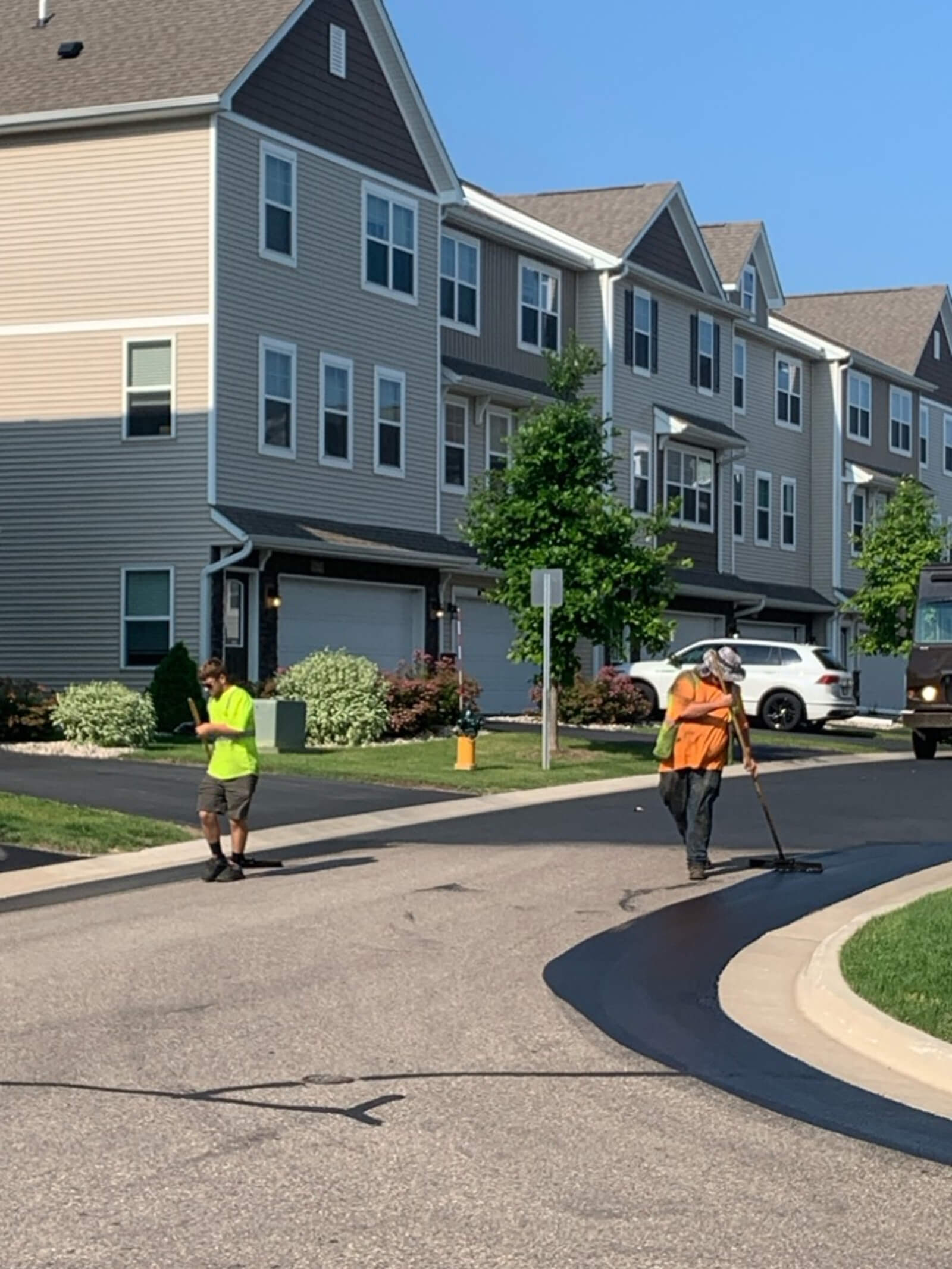 Two construction workers sealing a residential street driveway, using tools to apply a new layer of asphalt sealant on a bright, sunny day.