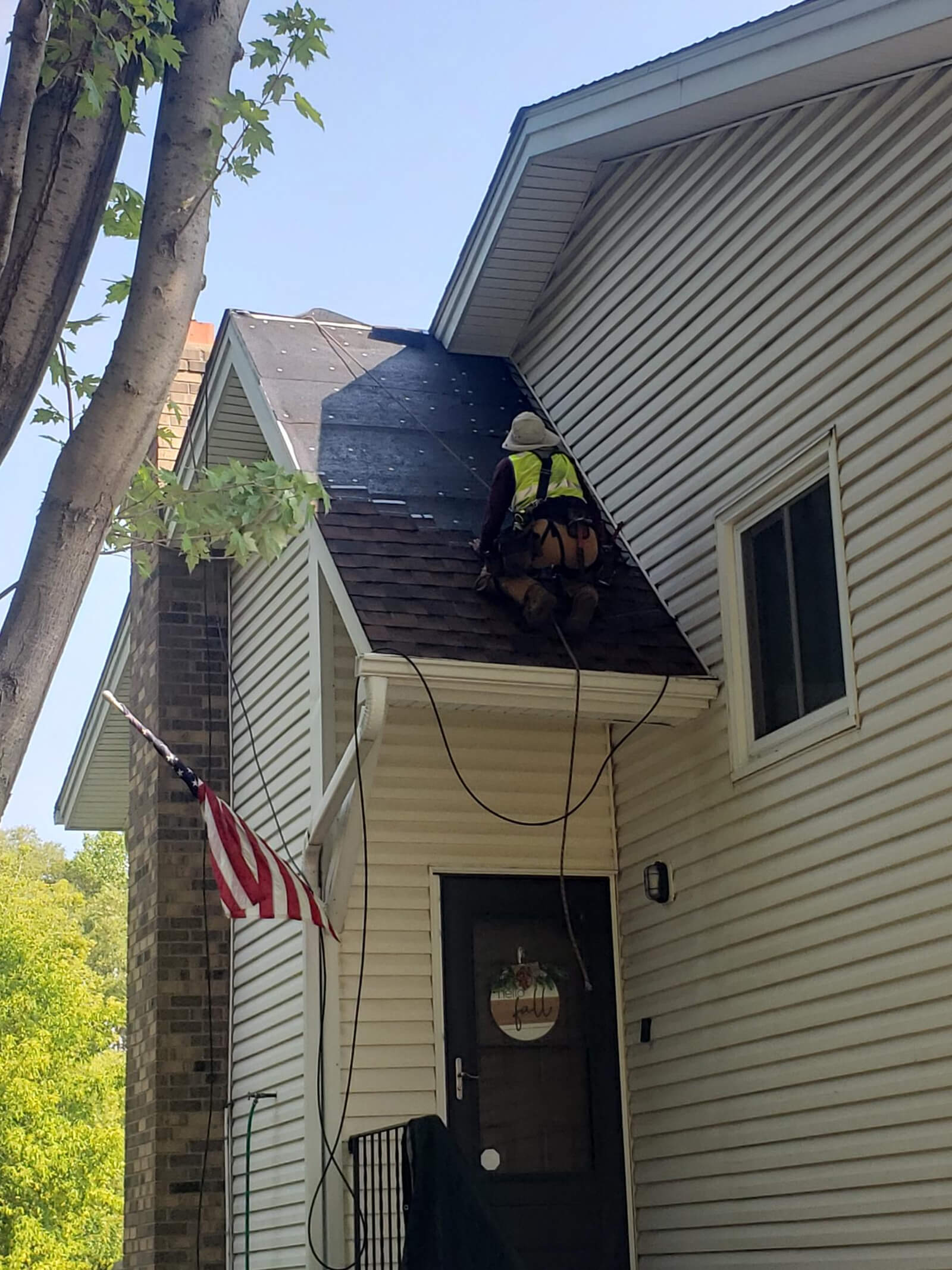 A construction worker in safety gear fixing shingles on a steep residential roof, with an American flag hanging by the front door.