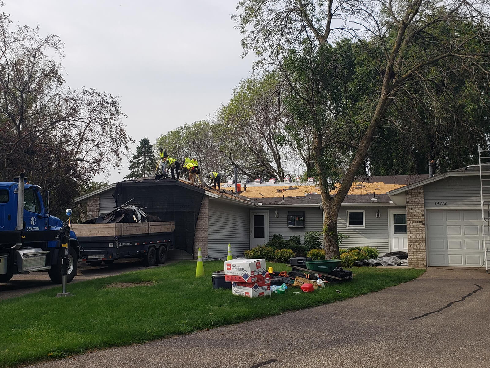 A group of roofers installing new shingles on a suburban home, surrounded by trucks, roofing materials, and tools.