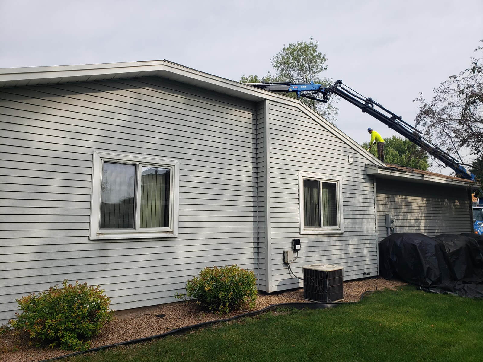 A construction worker on top of a house installing roofing materials with a crane assisting in lifting supplies.