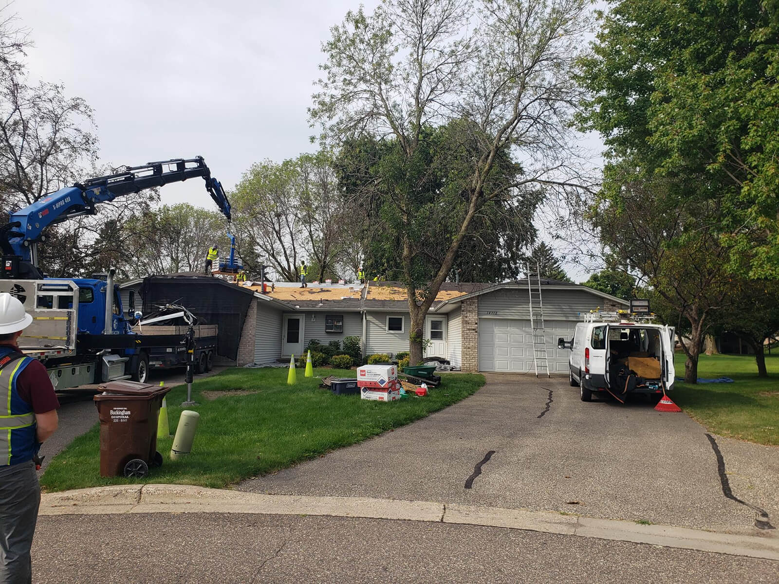 A team of roofers working to replace a roof on a suburban home, with vehicles and materials in the driveway.