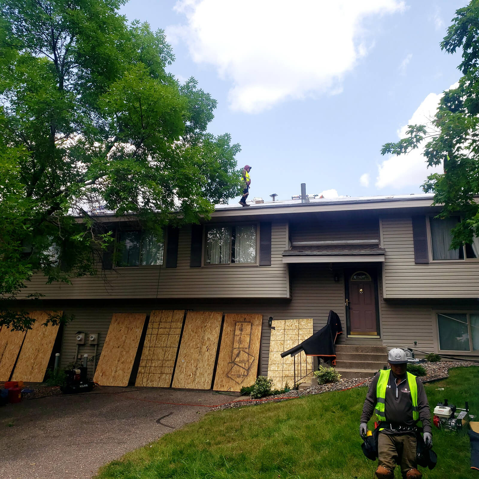 Workers in safety gear installing a new roof on a home. Wooden boards cover parts of the house, and roofing materials are placed around the property.