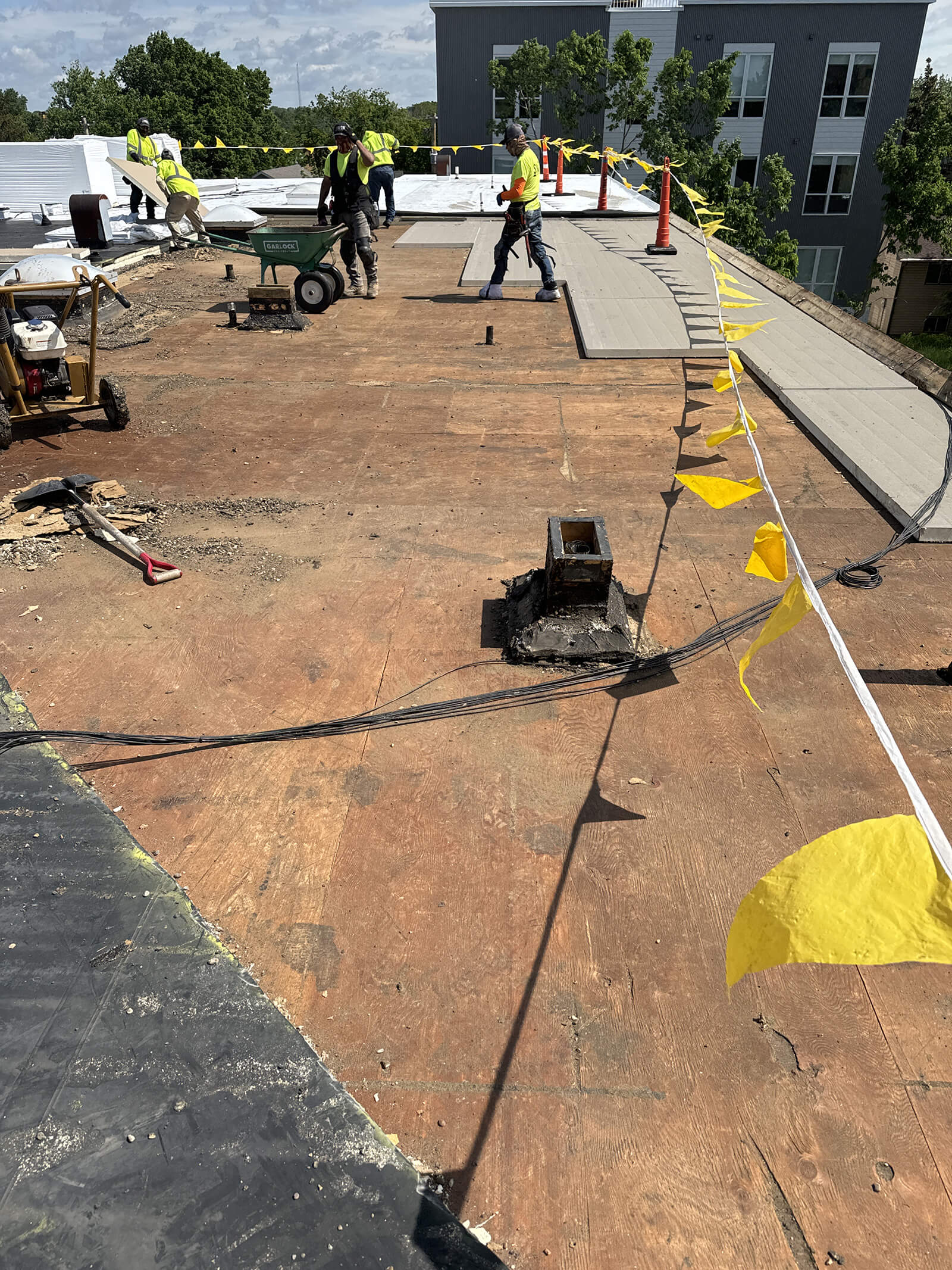 Construction workers preparing the roof for replacement, removing old materials and installing new panels, with caution tape marking the work area.