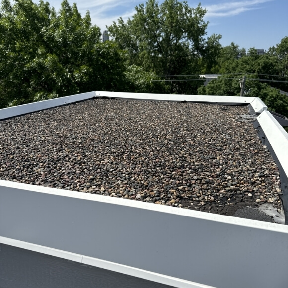 A flat roof covered with gravel, showing the protective edges of the roof and surrounded by trees in the background.