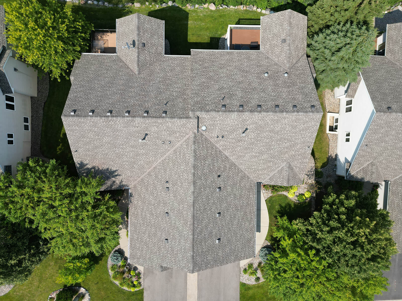 A drone-captured aerial view of a freshly installed residential roof, showing clean lines and shingles surrounded by trees and landscaping.