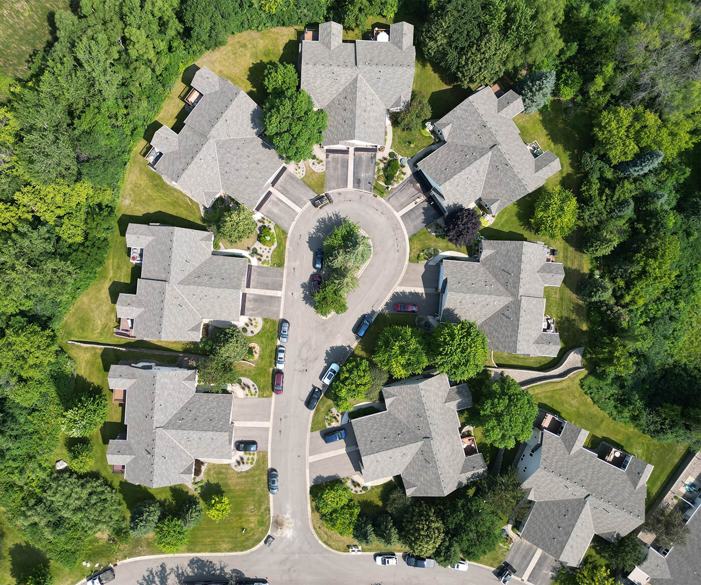 Aerial view of a residential cul-de-sac with houses surrounding a central roundabout, displaying well-maintained roofs, green lawns, and surrounding trees.