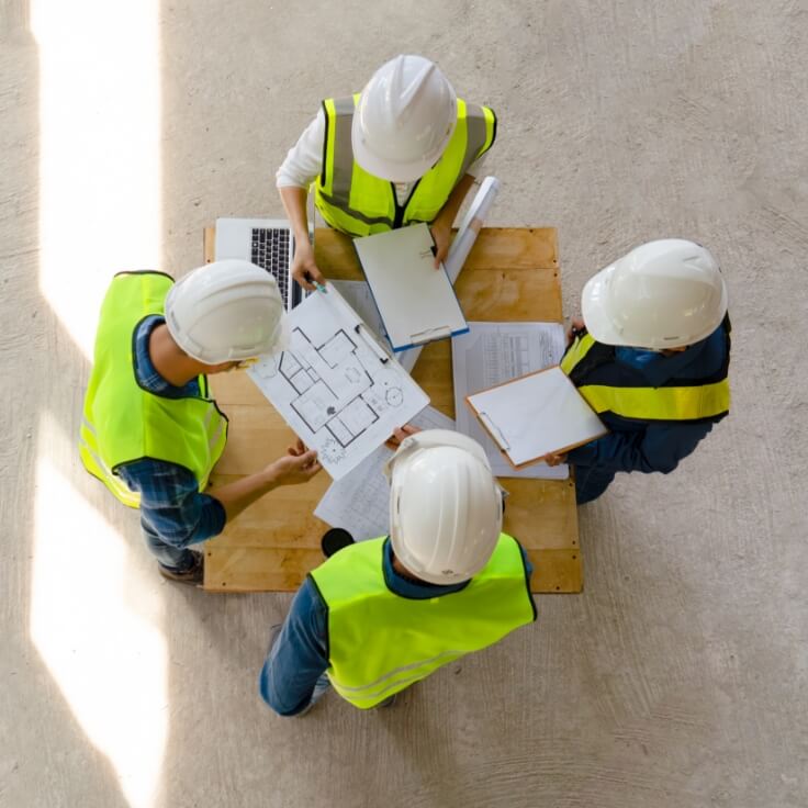 Group of construction workers wearing safety vests and helmets reviewing blueprints and documents on a table.